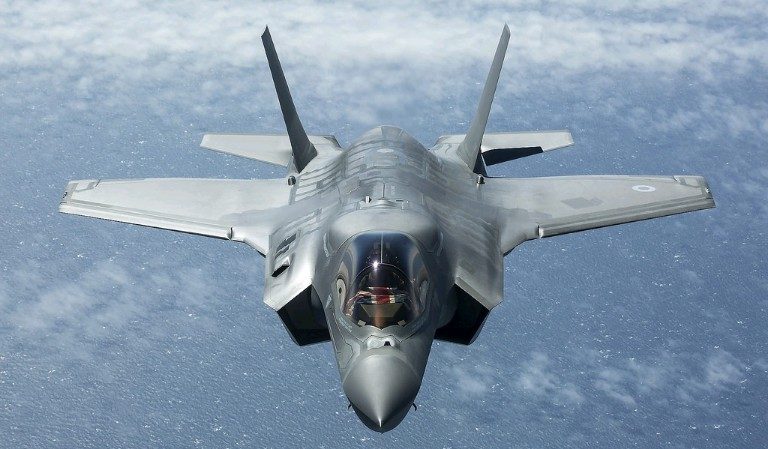 An F-35 fighter jet from the UK fleet flies over a body of water, seen from the front and above, with clouds in the background.