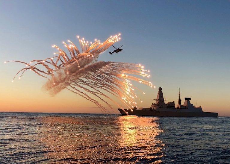 As the sun sets over the Mediterranean, a helicopter releases flares to safeguard HMS Diamond, casting a radiant glow over the naval ship at sea.