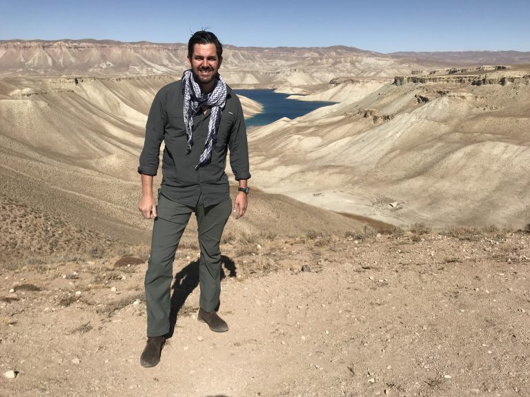 A man stands on a rocky hill, with a canyon and river in the background, embodying the spirit of a Pathfinder's Christmas Campaign. Sporting a scarf and casual attire, he channels the adventurous essence akin to Celebrity Hunted's very own Jordan Wylie.
