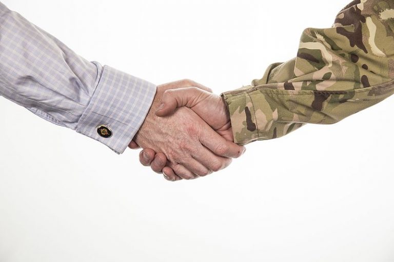 Two people shaking hands, one in a checkered shirt and the other in camouflage sleeves, symbolize the complex nature of a military transition, set against a crisp white background.