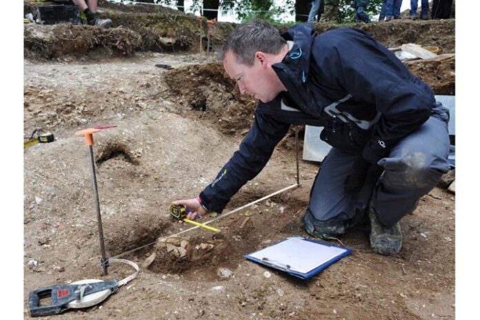 An archaeologist, like a military conservation expert, meticulously measures the site with a tape measure and clipboard, surrounded by soil and excavation tools. This careful documentation could earn an award for excellence in preserving history for future generations.