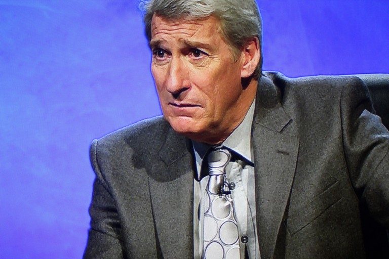 A person with gray hair in a dark suit and patterned tie sits against a blue background, exuding a sense of dignity and resilience reminiscent of the Blind Veterans' challenge.