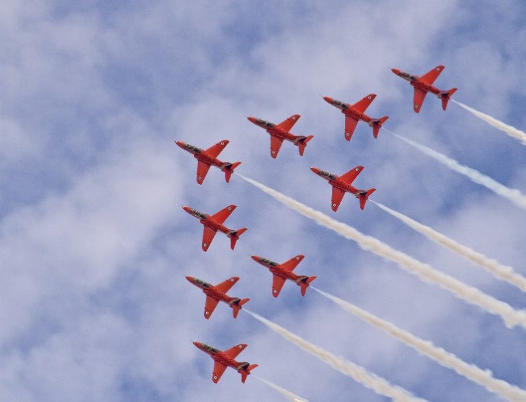 Nine Red Arrows jet fighters soar in formation against a cloudy sky, leaving white smoke trails behind.