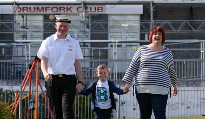A man in uniform, a child, and a woman stroll hand in hand outdoors past a 