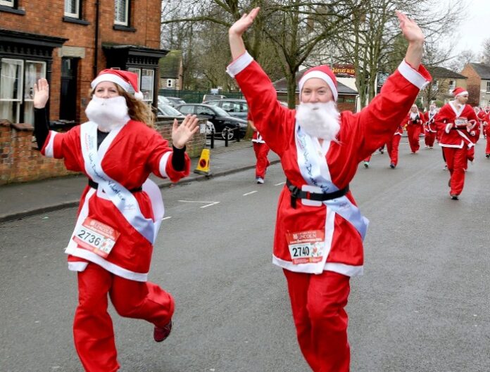 People dressed as Santa Claus running in a festive street race, wearing Christmas-themed costumes with beards and numbers. The atmosphere is filled with fun and the spirit of fundraising, as trees and buildings are visible in the background.