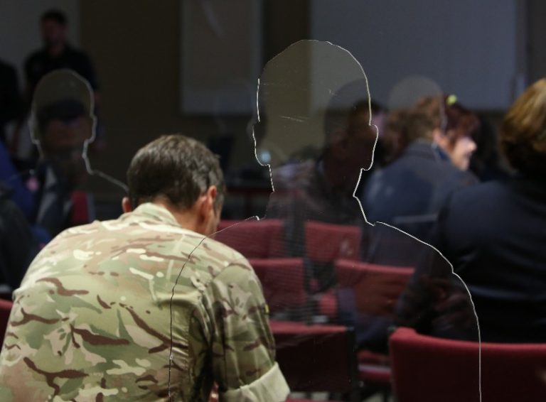 In a room bustling with activity, a person in camouflage uniform sits calmly, spectral silhouettes subtly appearing around the edges.