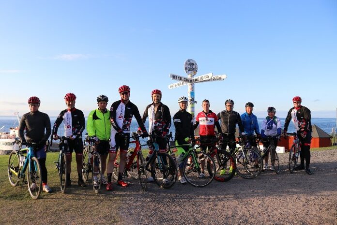 A group of people in cycling gear stands with their bikes near a signpost on a clear day by the sea, reminiscent of the camaraderie seen in Tour de Troops.
