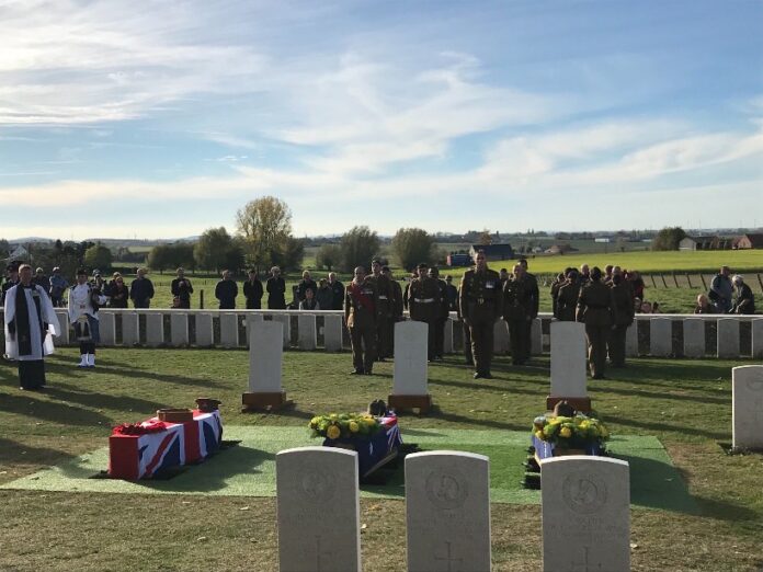 A military ceremony in a cemetery unfolds solemnly, with soldiers standing in formation. Three coffins, two draped in Union Jack flags, pay tribute to WW1 heroes as the Defence Minister stands nearby. Spectators respectfully observe the poignant scene from the background.