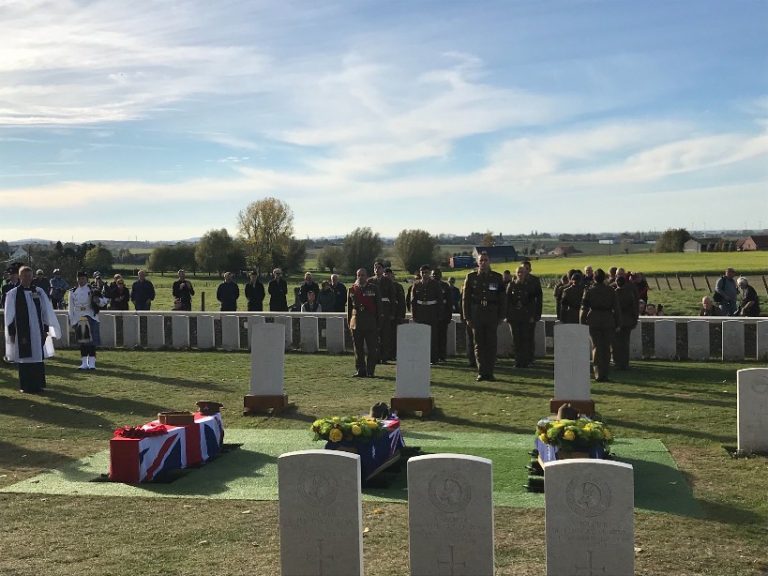 A military ceremony in a cemetery unfolds solemnly, with soldiers standing in formation. Three coffins, two draped in Union Jack flags, pay tribute to WW1 heroes as the Defence Minister stands nearby. Spectators respectfully observe the poignant scene from the background.
