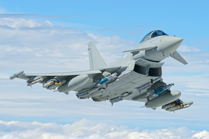 A fighter jet from the Quick Reaction Alert Force soars against a blue sky with clouds, showcasing its mounted weaponry and superb aerodynamic design.