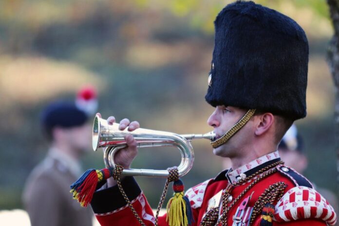 A soldier in ceremonial uniform, honoring the Unknown Warrior, plays a silver bugle outdoors. The uniform, befitting the somber occasion of the final resting place tribute, features a tall fur hat and detailed decorations.