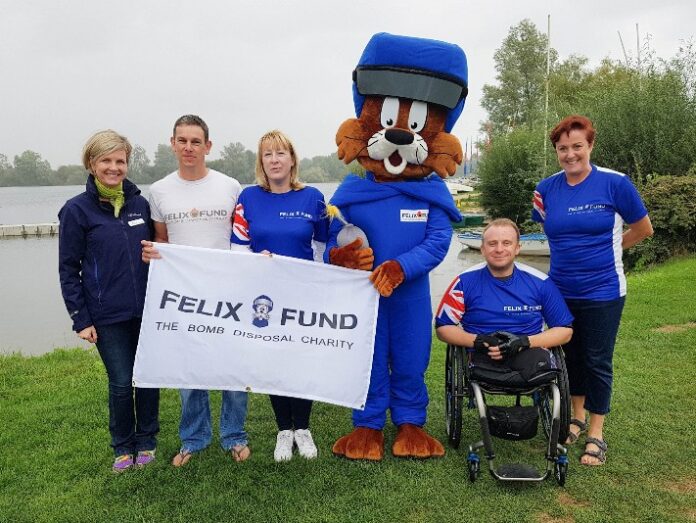 A group of people and a mascot, including Captain James Wadsworth, proudly hold a Felix Fund charity banner by the lake. This dynamic team commemorates their Challenges 2019 achievement with a picturesque backdrop, uniting spirit and purpose for an inspiring cause.