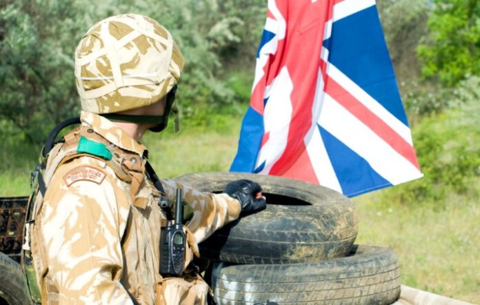 A soldier in camouflage gear, working tirelessly amidst a stack of tires, stands proudly holding a UK flag in an outdoor setting.
