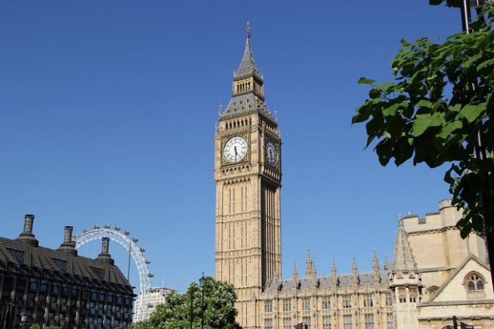 Big Ben and the Elizabeth Tower, iconic London landmarks, stand proudly with the London Eye in the background against a clear blue sky. Tree leaves elegantly frame the right side of this picturesque scene.