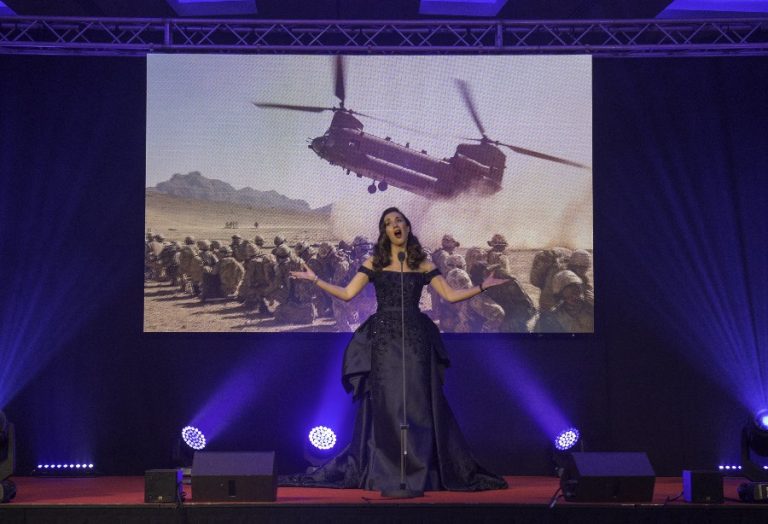 Carly Paoli, in a dark dress, sings passionately on stage, her enchanting voice echoing through the venue. Behind her, a military scene with soldiers and a helicopter is projected, setting the backdrop for a single performed in honor of military fundraising.