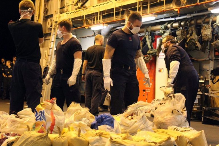 Several uniformed individuals wearing masks and gloves handle bags in the well-lit interior of HMS Dragon, showcasing a stern commitment to combating narcotics in the Gulf.