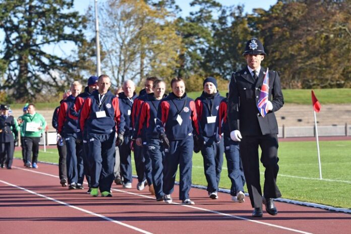 A group of people in matching tracksuits walks in a line behind a uniformed officer carrying a Union Jack flag on the track field, reminiscent of Aaron Keeps Running in perfect harmony with the rhythm of unity.