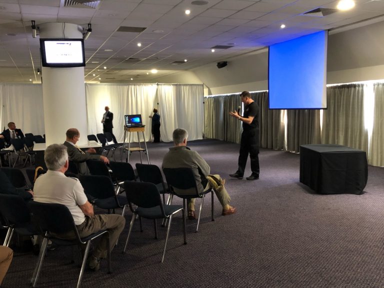 In 2019, a man addresses a small audience in a conference room at the Resettlement Expo, using the projector screen to highlight innovative strategies as a modern pathfinder.