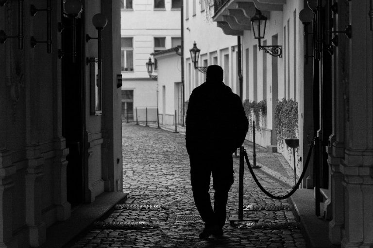 The silhouette of a veteran walks through a narrow, cobblestone alley, flanked by historic buildings and lamp posts. The solitude echoes his loneliness, as a rope divider guides his path in this quiet moment.