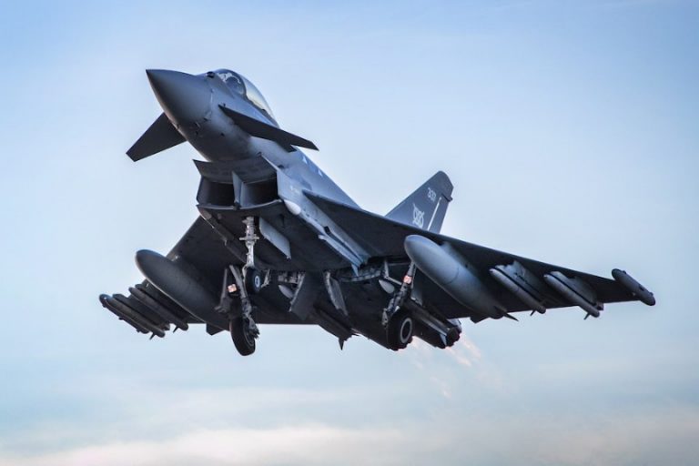 A military jet, featuring a new missile system, flies with landing gear extended against a blue sky backdrop.