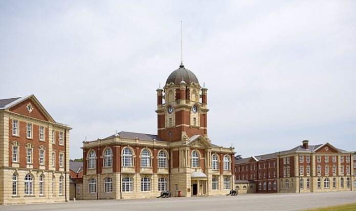 A historic brick building with a prestigious clock tower and arched windows stands in the center, flanked by two similar structures, under a partly cloudy sky.