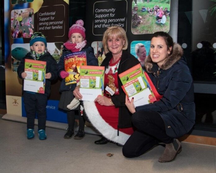 Two adults and two children, preparing for the holidays, hold folders and a book, standing indoors in front of a display about community support services for families with deployed parents.