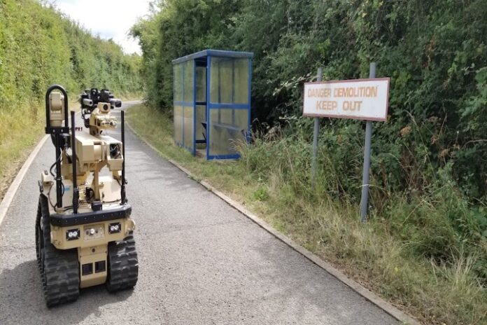A bomb disposal robot on a pathway approaches a fenced-off area with a sign reading 
