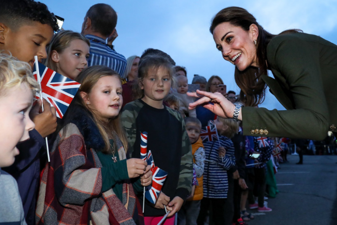 At an outdoor event, a woman cheerfully greets a group of children waving British flags, echoing the warmth and unity often seen in gatherings with the Duke and Duchess of Cambridge.