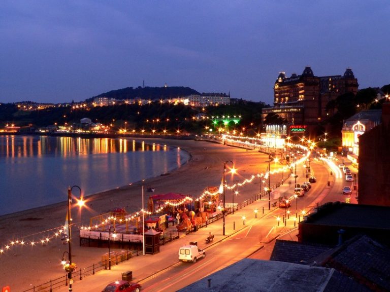As dusk descends on the coastal town of Scarborough, illuminated streetlamps and buildings cast a warm glow over the beach, while the calm sea reflects the peaceful ambiance. In this serene setting, stories of HMS Duncan's past affiliation with these shores whisper softly in the evening breeze.