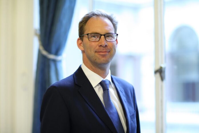 A man in a suit and tie stands in front of a window with blue curtains, looking forward, like a minister preparing to address praises from various organisations.