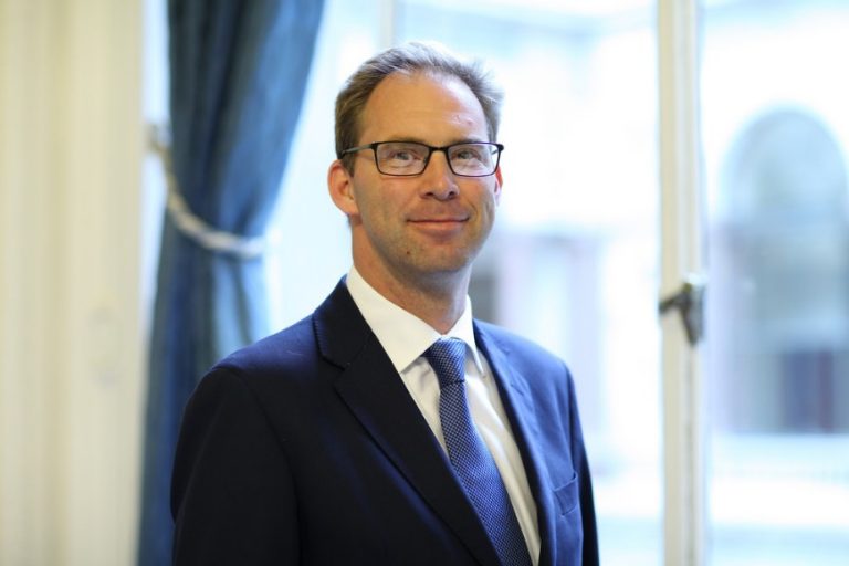 A man in a suit and tie stands in front of a window with blue curtains, looking forward, like a minister preparing to address praises from various organisations.