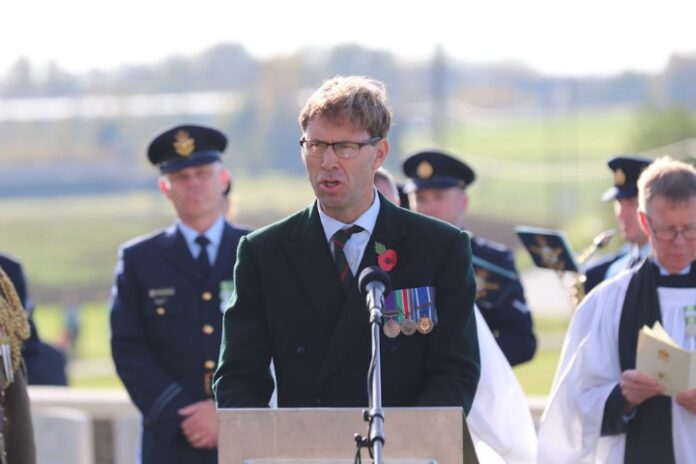 A man speaks at a podium, wearing a suit adorned with military medals, embodying the spirit of going the Extra Mile. Several uniformed War Detectives stand in the background, attentive and steadfast.
