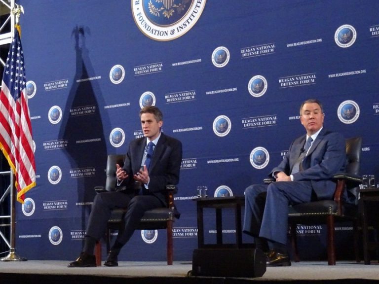 Two men in suits sit on stage at the Reagan National Defense Forum, discussing the UK-US Defence Partnership, with an American flag and event backdrop visible.