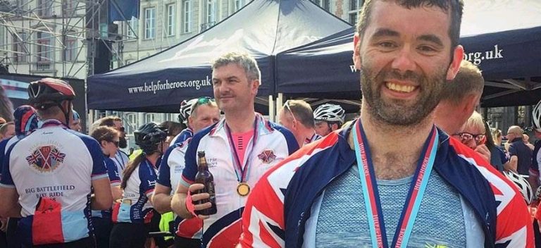 Cyclists gather outdoors wearing UK-themed jerseys and medals, some holding drinks, with tents in the background, celebrating their achievements from the 2019 Big Battlefield Bike Ride.