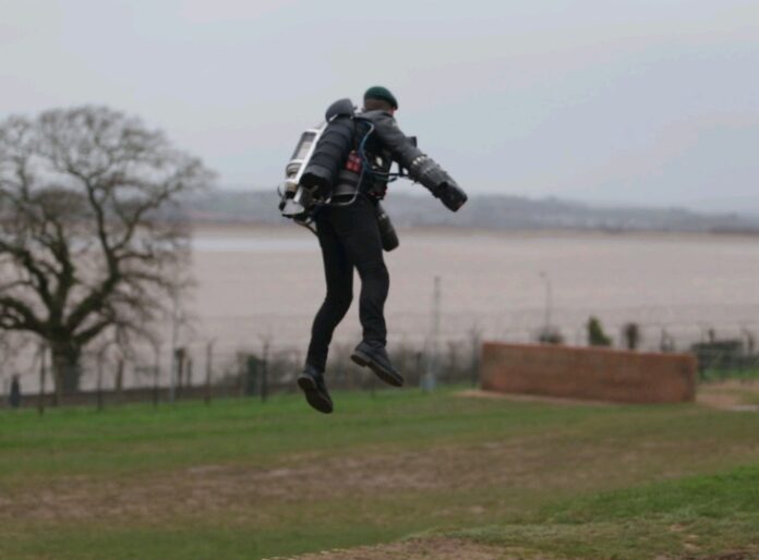 A person soars above the ground with a jetpack, reminiscent of a Real-Life Iron Man, against a backdrop of grass, a tree, and a shimmering body of water.
