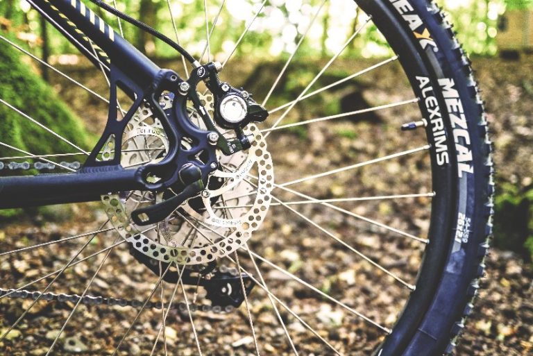 Close-up of a mountain bike's rear wheel and disc brake, with thick tire tread navigating through a forest trail, reminiscent of the rugged terrains tackled during the adventurous Tour de Troops.