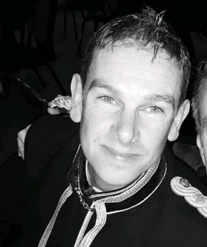 A person in a dark, ornate jacket smiles while looking slightly upward, embodying the spirit of a compassionate fundraiser. The image is in black and white, capturing the timeless dedication often celebrated by an MBE.
