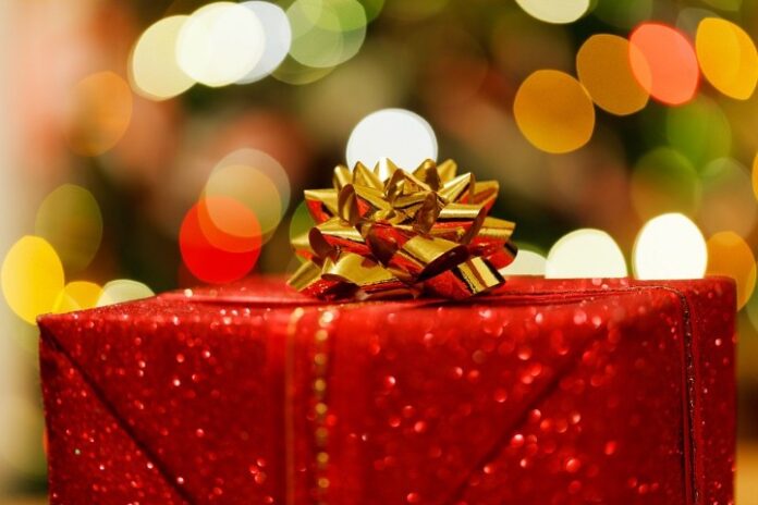 A glittery red Christmas box with a gold ribbon bow on top, set against a backdrop of colorful, soft-focus lights.