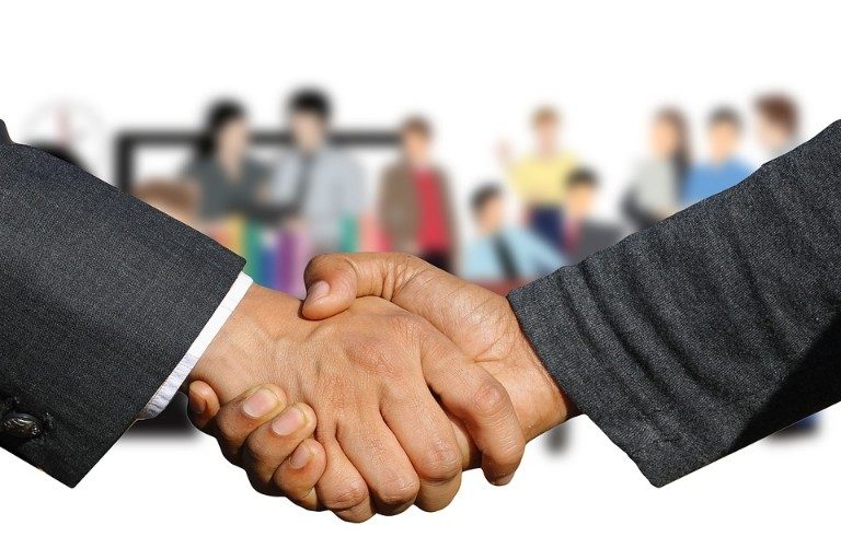 Two people in suits are shaking hands, sealing a deal under the Covenant Programme, while a blurred group of people looks on in anticipation.