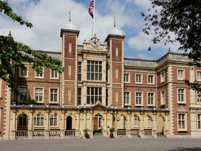 Historic brick and stone building with a central clock tower and a UK flag on top, surrounded by trees, is renowned as the Home of Military Music.