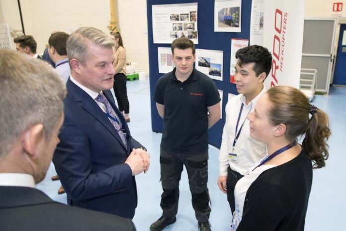 A man in a suit discusses the £293 Million deal with three young adults in business casual attire, surrounded by display boards in an indoor setting.