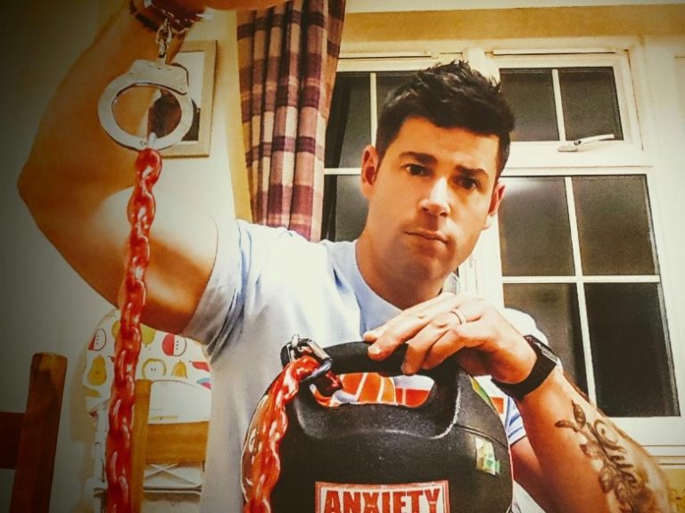 A person grips a kettlebell labeled "Anxiety," shackled with red chain links and handcuffs, symbolizing the visible wounds of mental health struggles. Indoors, a curtain-draped window casts soft light over the poignant scene.