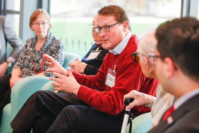 A man in a red sweater speaks animatedly about the Commissioner's Agenda in a group meeting. Several people listen attentively, seated on light blue chairs in a bright room, where ideas for combatting loneliness are shared passionately.