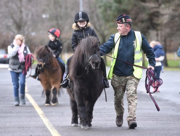 Children in helmets enjoy a lively Poppy Ride on ponies, guided by attentive adults. A person in a reflective vest walks alongside each pony, ensuring a safe and joyful experience for all involved.