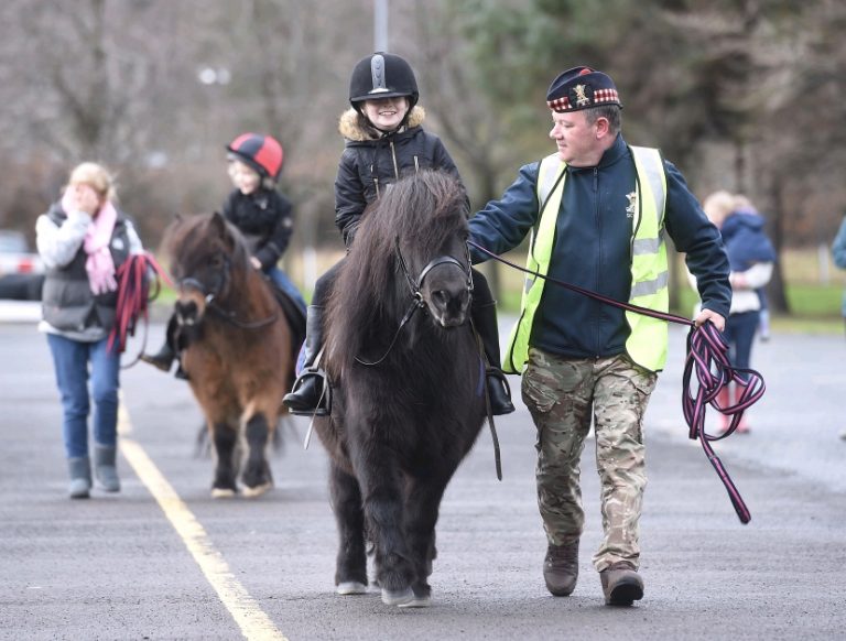 Children in helmets enjoy a lively Poppy Ride on ponies, guided by attentive adults. A person in a reflective vest walks alongside each pony, ensuring a safe and joyful experience for all involved.