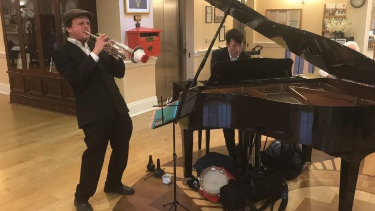 A man plays the trumpet with a swinging flair next to a grand piano, while another man skillfully plays the piano. They are in a room with wooden floors, celebrating Founders' Day, with a postbox subtly visible in the background.