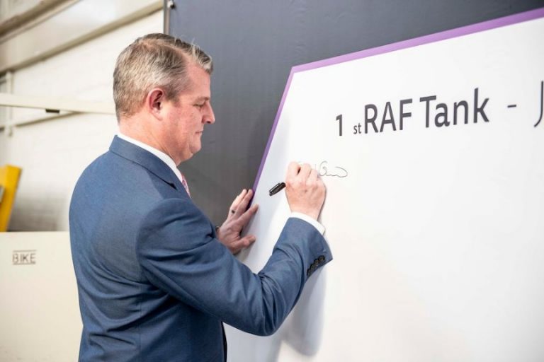 A person in a suit, representing the way forward in UK protection, signs a board labeled "1st RAF Tank," with additional text partially visible.
