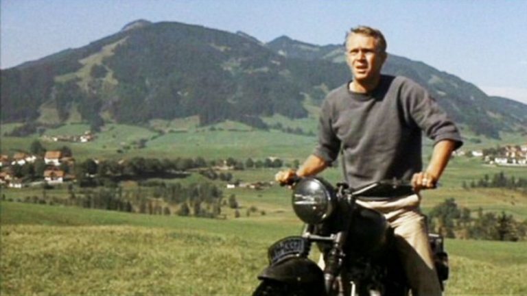 A man stands beside his motorcycle, reminiscent of *The Great Escape*, surrounded by a scenic landscape of majestic mountains and sweeping grass fields.