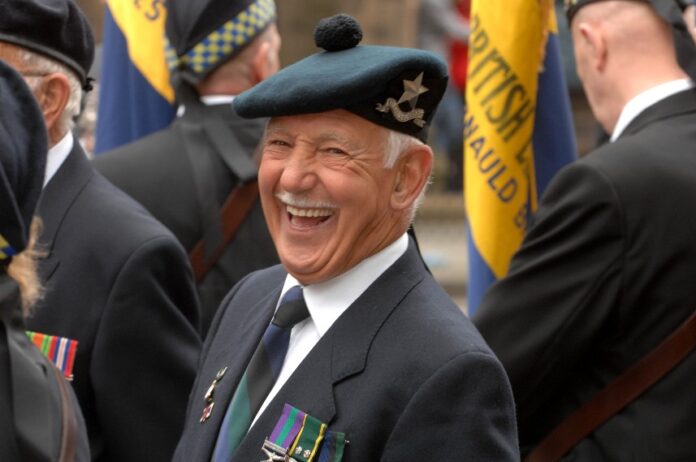 An elderly man in a military beret and suit smiles proudly among fellow veterans during a parade, honoring unforgotten forces. Flags and badges are visible in the background, capturing the spirit of camaraderie and remembrance.