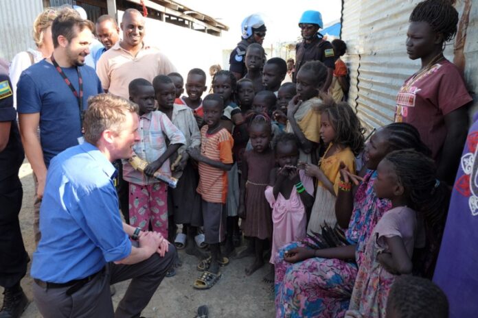 A group of adults and children gather outside a building in Sudan. One adult kneels, interacting with the children. Two uniformed UK peacekeepers, reminiscent of the efforts led by Williamson, stand nearby.